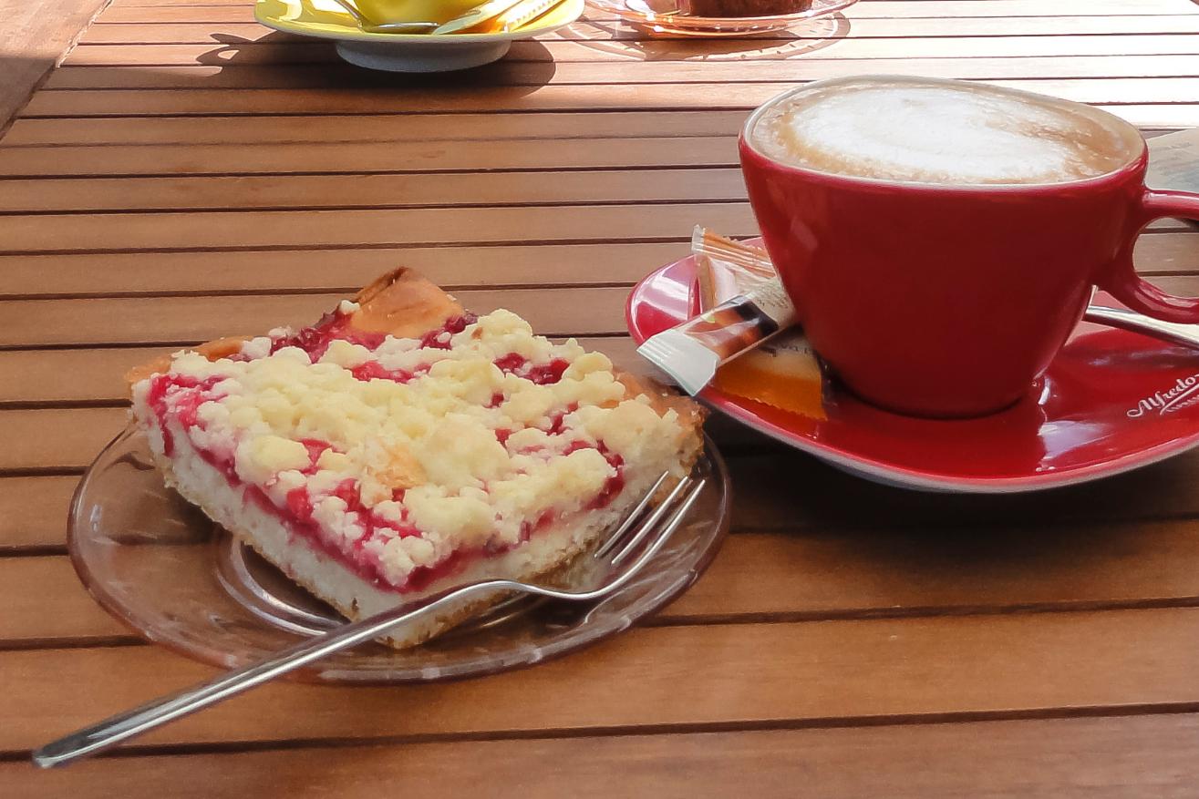Man sieht ein Stück Streuselkuchen auf einem roten Teller, rechts daneben eine rote Tasse mit Kaffee auf einer Untertasse