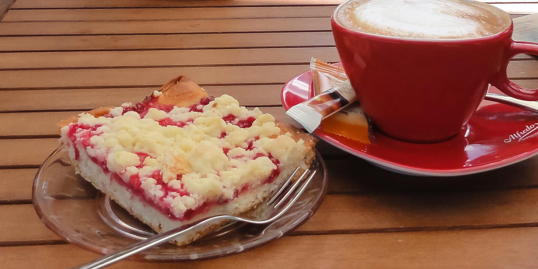 Man sieht ein Stück Streuselkuchen auf einem roten Teller, rechts daneben eine rote Tasse mit Kaffee auf einer Untertasse