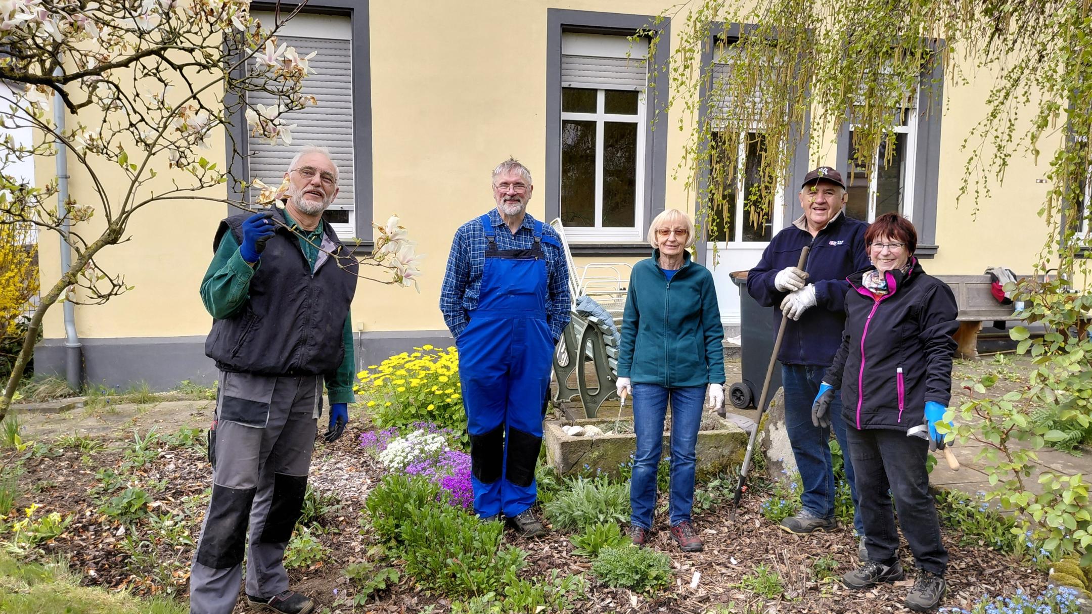 Man sieht eine Gruppe Männer und Frauen vor einem gelb gestrichenen Gebäude, in einem Garten stehen