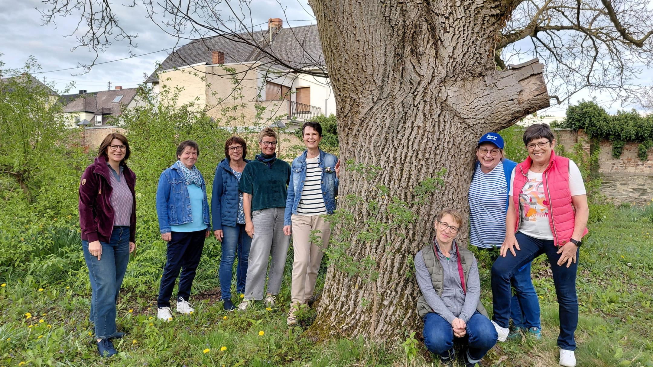 Man sieht eine Gruppe Frauen, die in einem Garten rund um einen Baum, auf einer grünen Wiese stehen