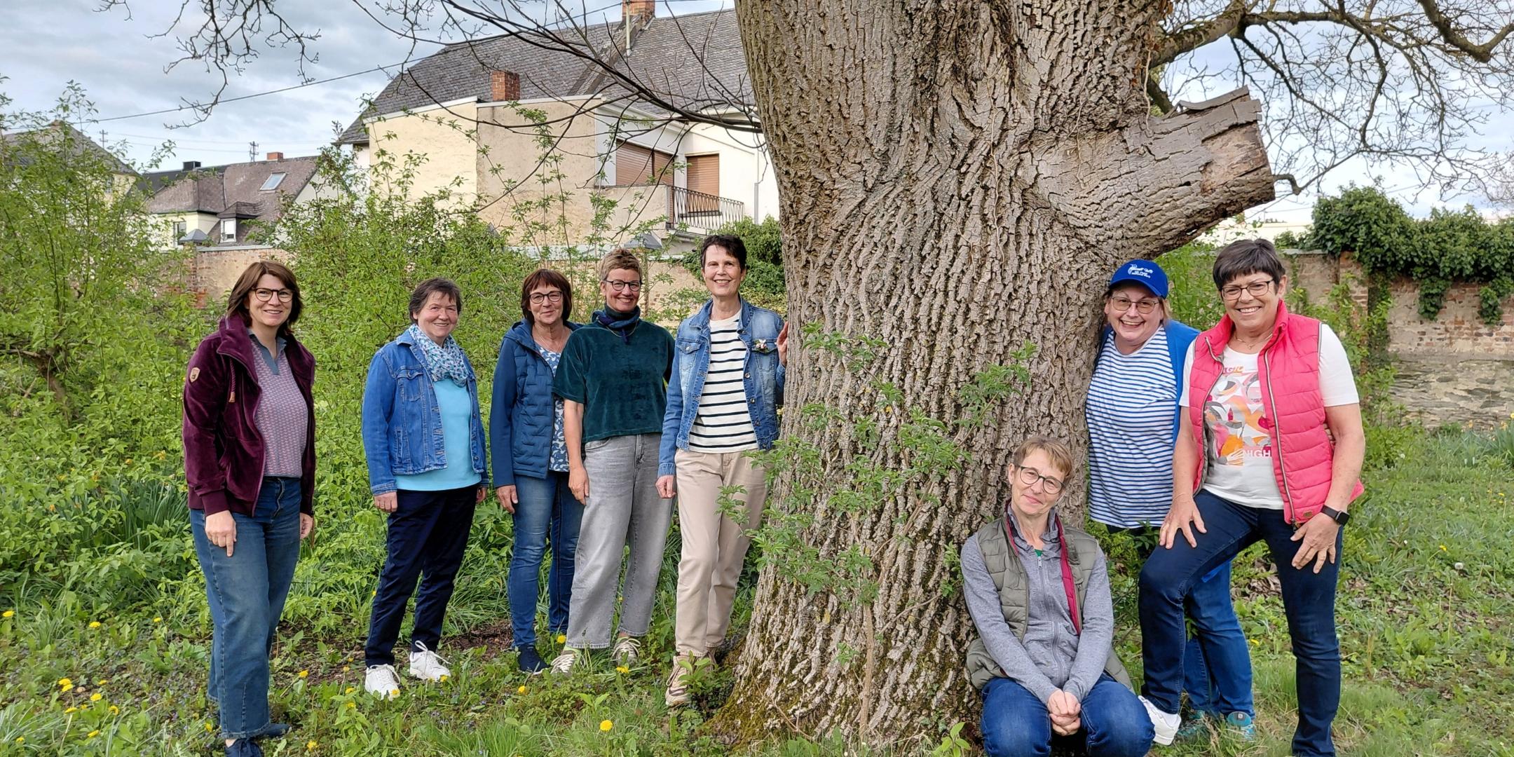 Man sieht eine Gruppe Frauen, die in einem Garten rund um einen Baum, auf einer grünen Wiese stehen