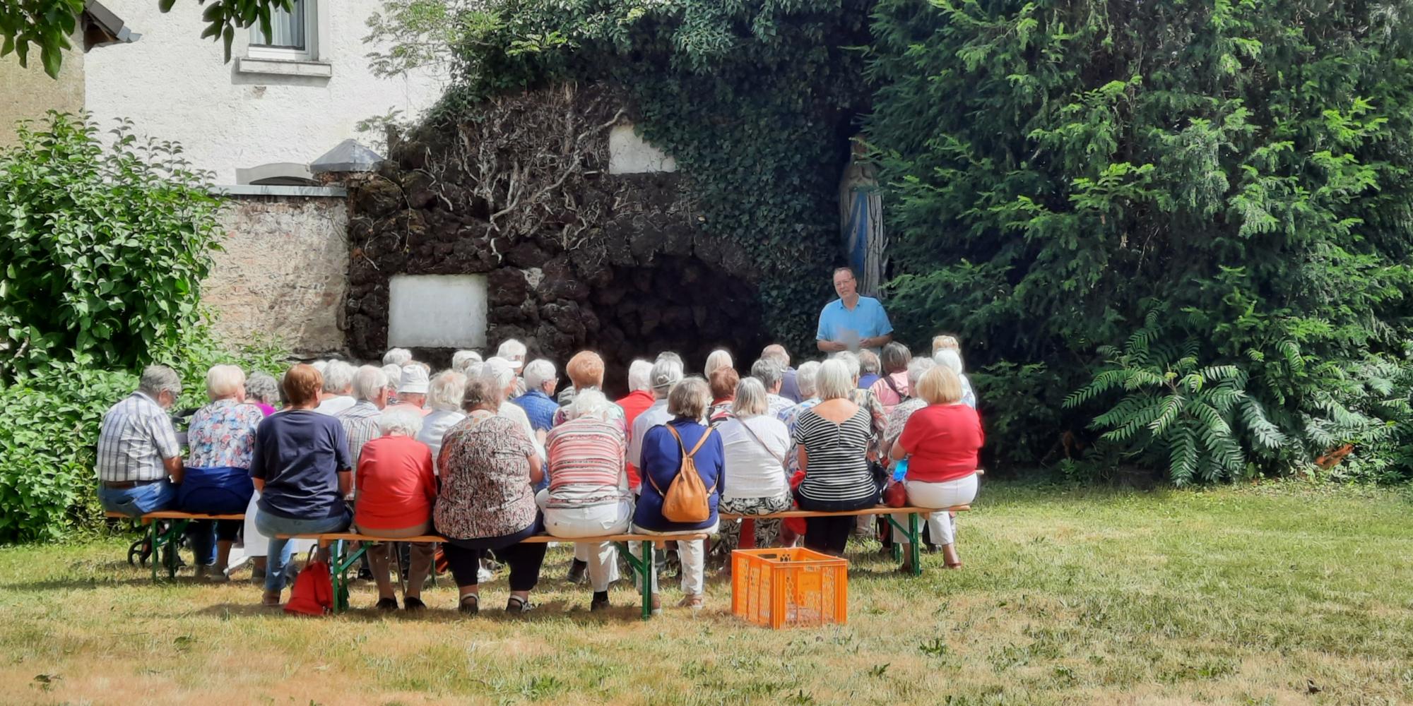 Man sieht eine Gruppe Menschen auf Bänken in einem Garten vor einer Mariengrotte sitzend, davor ein Mann, der etwas erzählt. (c) Privat Man sieht eine Gruppe Menschen auf Bänken in einem Garten vor einer Mariengrotte sitzend, davor ein Mann, der etwas erzählt.