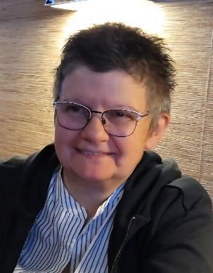 Man sieht eine Frau mit blau gestreifter Bluse, schwarzer Jacke, schwarzen Haaren und Brille, im Hintergrund das Meer