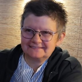 Man sieht eine Frau mit blau gestreifter Bluse, schwarzer Jacke, schwarzen Haaren und Brille, im Hintergrund das Meer
