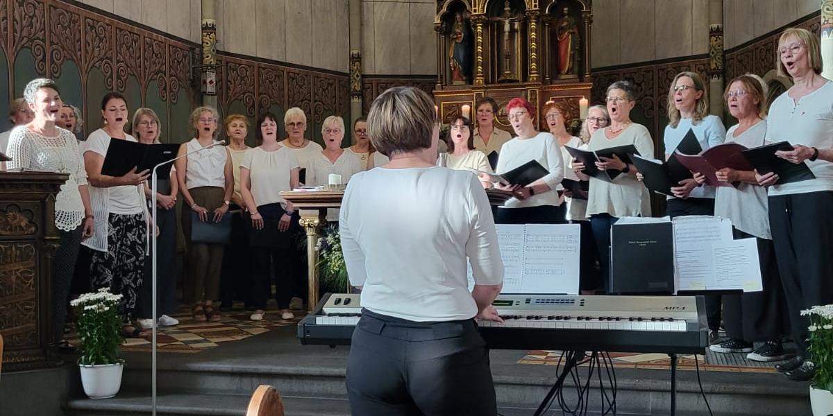Man sieht Frauen mit Notenbüchern in weißen Blusen und schwarzen Hosen in der Apsis der Kirche in Rüber singen, davor die Chorleiterin (c) Privat Man sieht Frauen mit Notenbüchern in weißen Blusen und schwarzen Hosen in der Apsis der Kirche in Rüber singen, davor die Chorleiterin