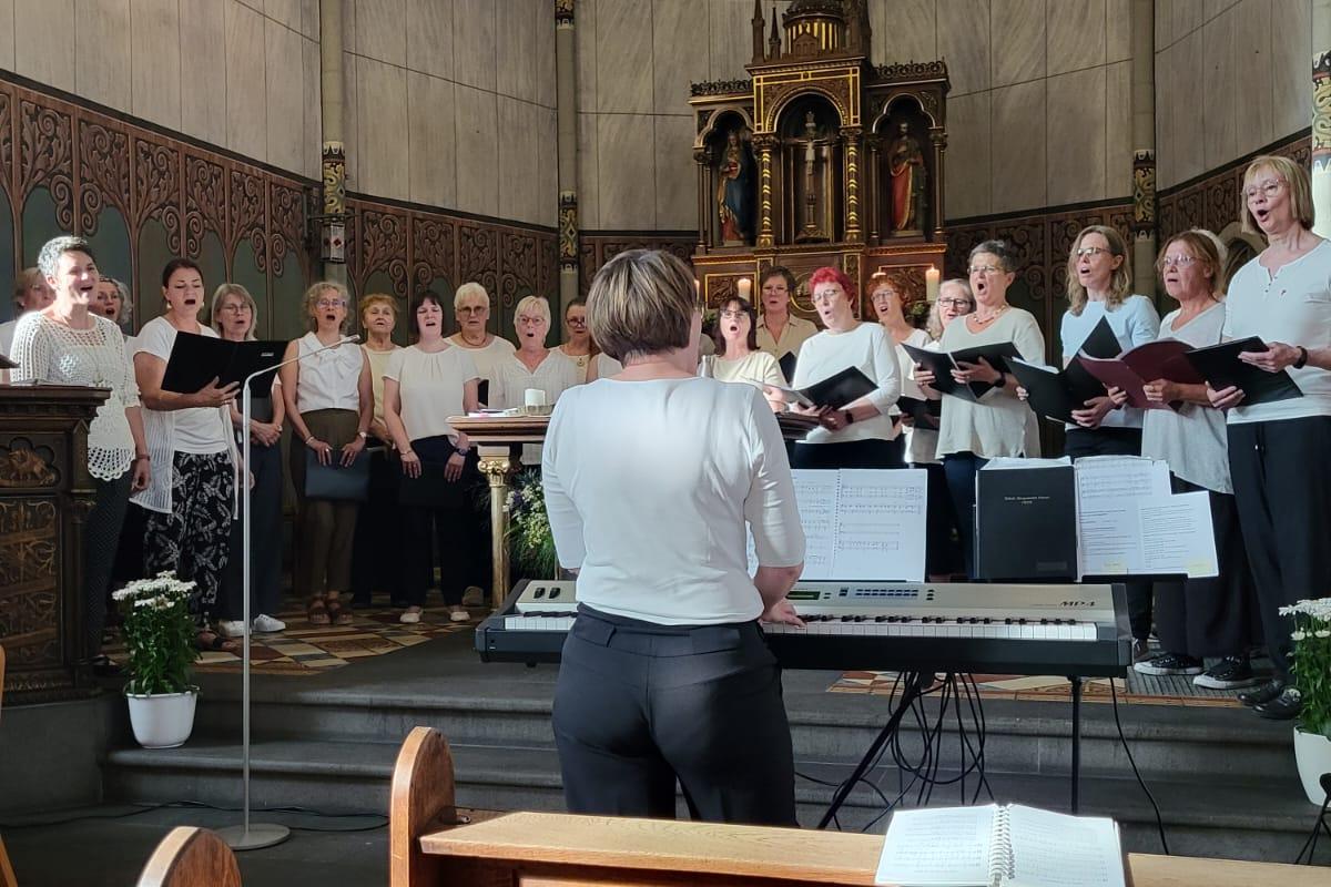 Man sieht Frauen mit Notenbüchern in weißen Blusen und schwarzen Hosen in der Apsis der Kirche in Rüber singen, davor die Chorleiterin
