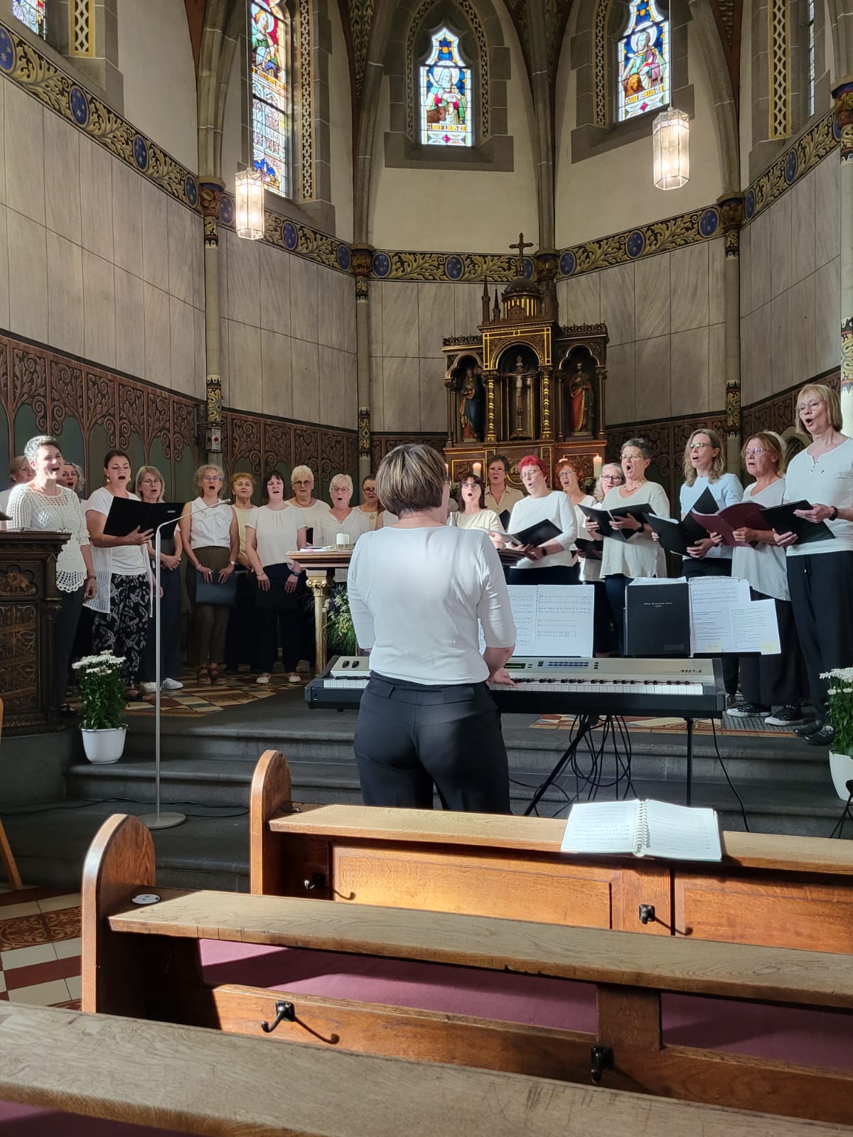 Man sieht Frauen mit Notenbüchern in weißen Blusen und schwarzen Hosen in der Apsis der Kirche in Rüber singen, davor die Chorleiterin