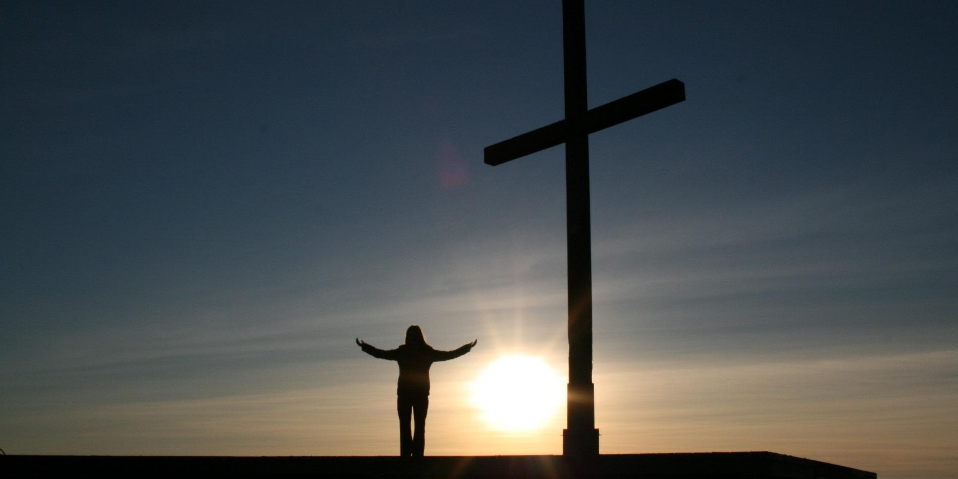 Man sieht vor untergehender Sonne im Gegenlicht eine Frau, die Hände zum Gebet geöffnet, neben einem Kreuz.