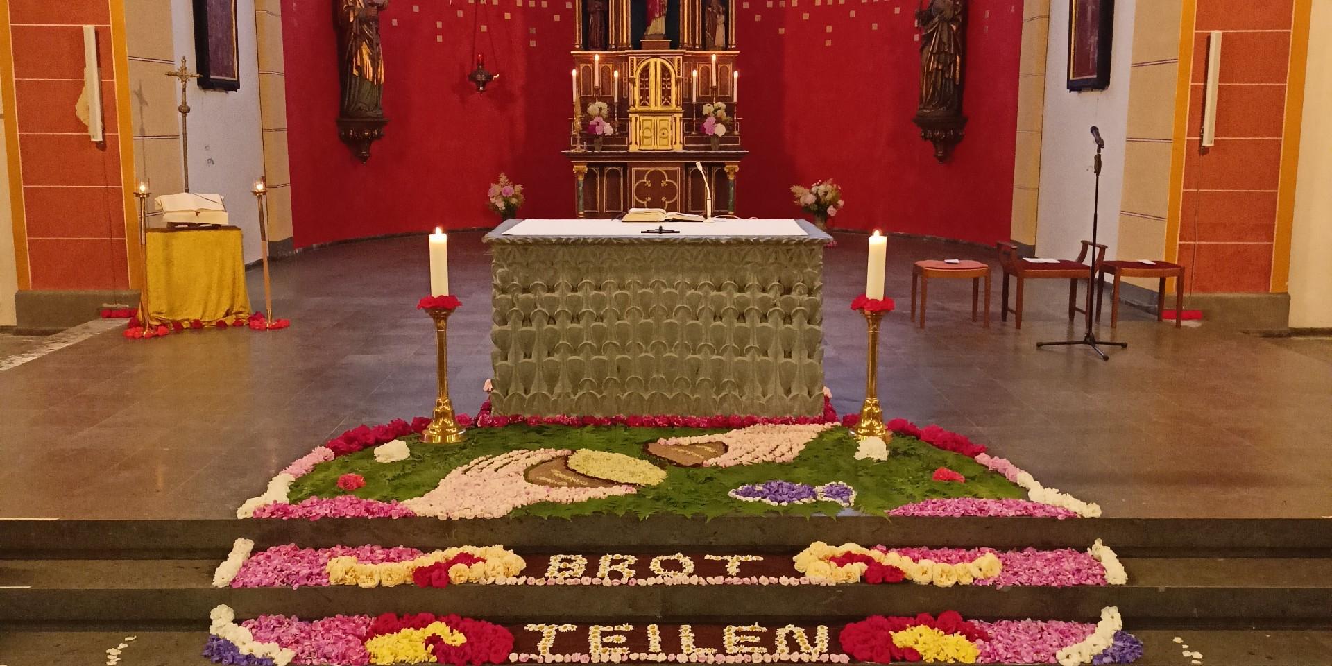 Man sieht einen bunten Blumenteppich vor einem Altar - mit dem Schriftzug 'Brot teilen'