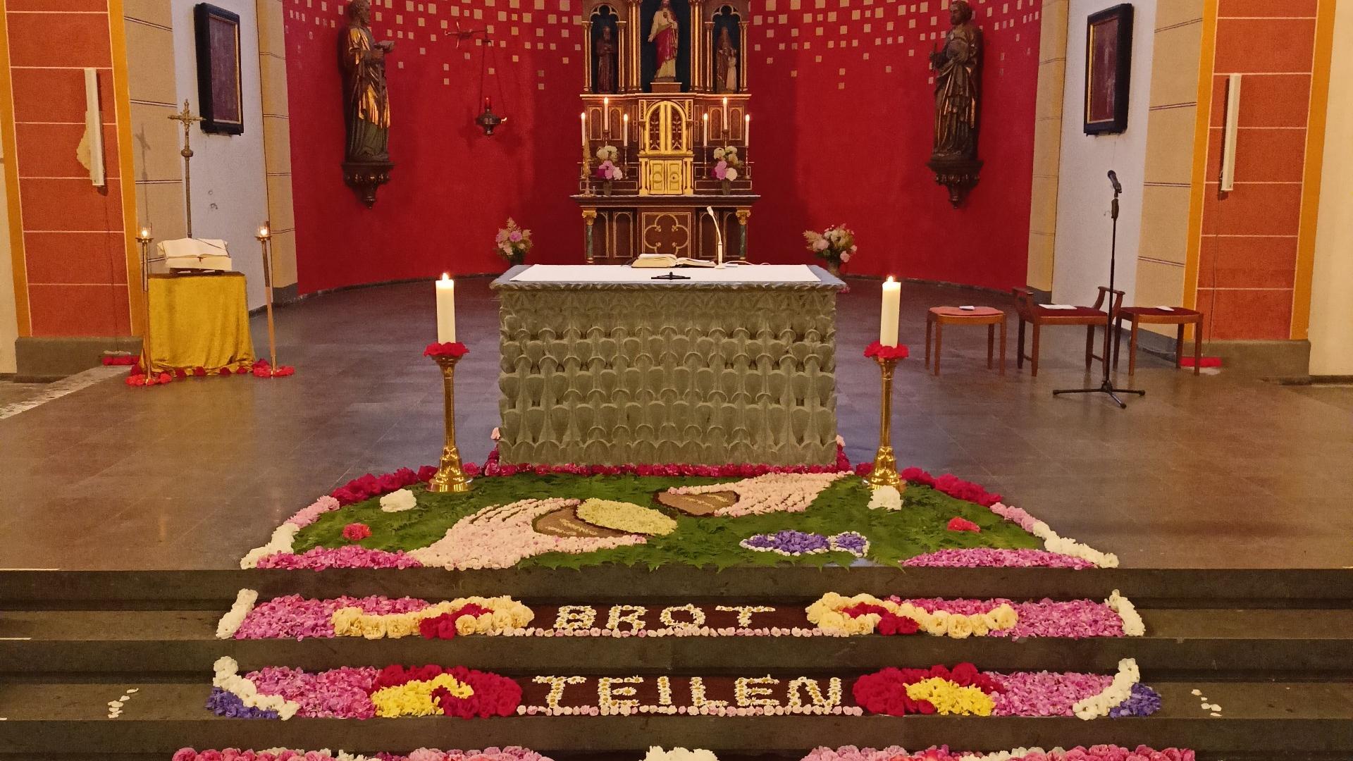 Man sieht einen bunten Blumenteppich vor einem Altar - mit dem Schriftzug 'Brot teilen'