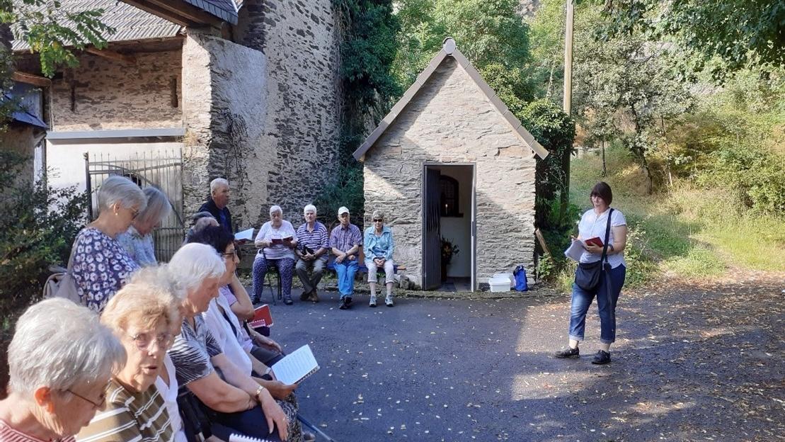 Man sieht eine Frau, die vor einer Gruppe Menschen , die auf Bänken an einer Kapelle sitzen, einen Text vortragen