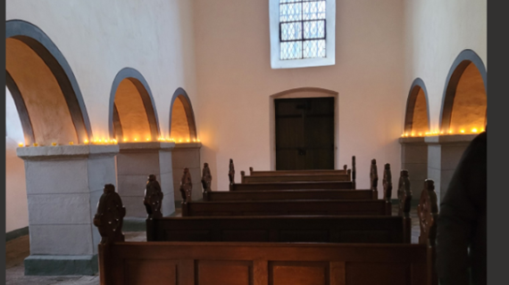 Man sieht den weiß gestrichenen Innenraum der Georgskapelle, auf den Simsen der Pfeiler stehen brennende Kerzen, welche die Kapelle erhellen