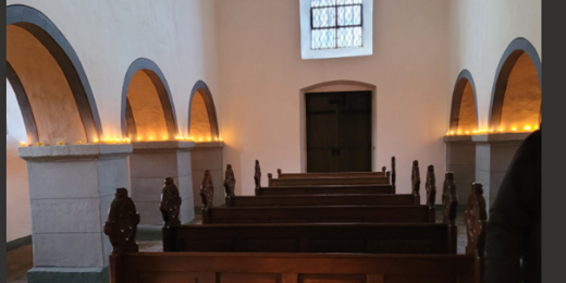 Man sieht den weiß gestrichenen Innenraum der Georgskapelle, auf den Simsen der Pfeiler stehen brennende Kerzen, welche die Kapelle erhellen