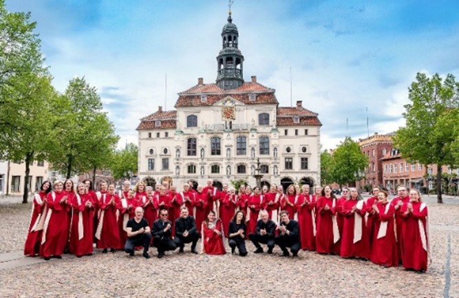 Man sieht eine Gruppe von Menschen in roten Talare gekleidet und davor ein Konzertensemble in schwarz gekleidet vor einen weißen im Renaissance-Stil gehaltenen Gebäude stehen