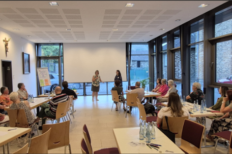 Man sieht zwei Frauen im Vordergrund, die eine Veranstaltung moderieren und Menschen, die an Tischen sitzend, der Veranstaltung lauschen