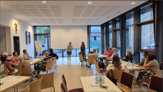 Man sieht zwei Frauen im Vordergrund, die eine Veranstaltung moderieren und Menschen, die an Tischen sitzend, der Veranstaltung lauschen (c) Pastoraler Raum Maifeld-Untermosel/Privat Man sieht zwei Frauen im Vordergrund, die eine Veranstaltung moderieren und Menschen, die an Tischen sitzend, der Veranstaltung lauschen