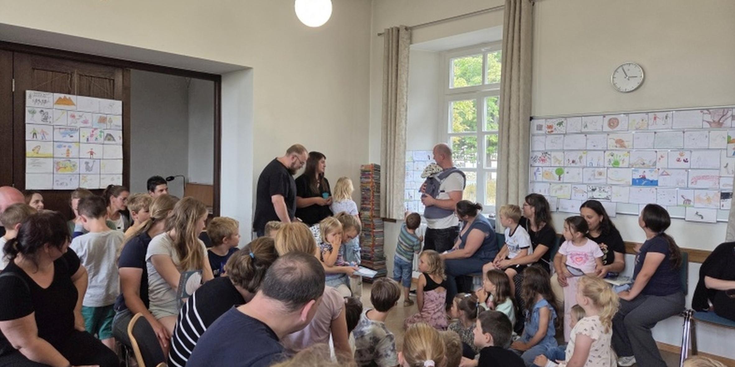 KÖB Münstermaifeld Abschlussfest Lesesommer (1) (c) Privat Mans sieht viele Eltern mit Kinder in einem hellgelb gestrichenen Raum sitzend, die auf drei stehende Personen achten