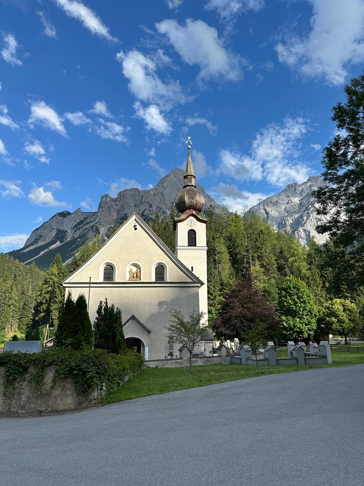Man sieht eine Kirche in einer Bergregion, vor einem Berg und blauem, mit weißen Wolken geschmückten Himmel. Der Kirchturm ist mit einem Zwiebeldach versehen