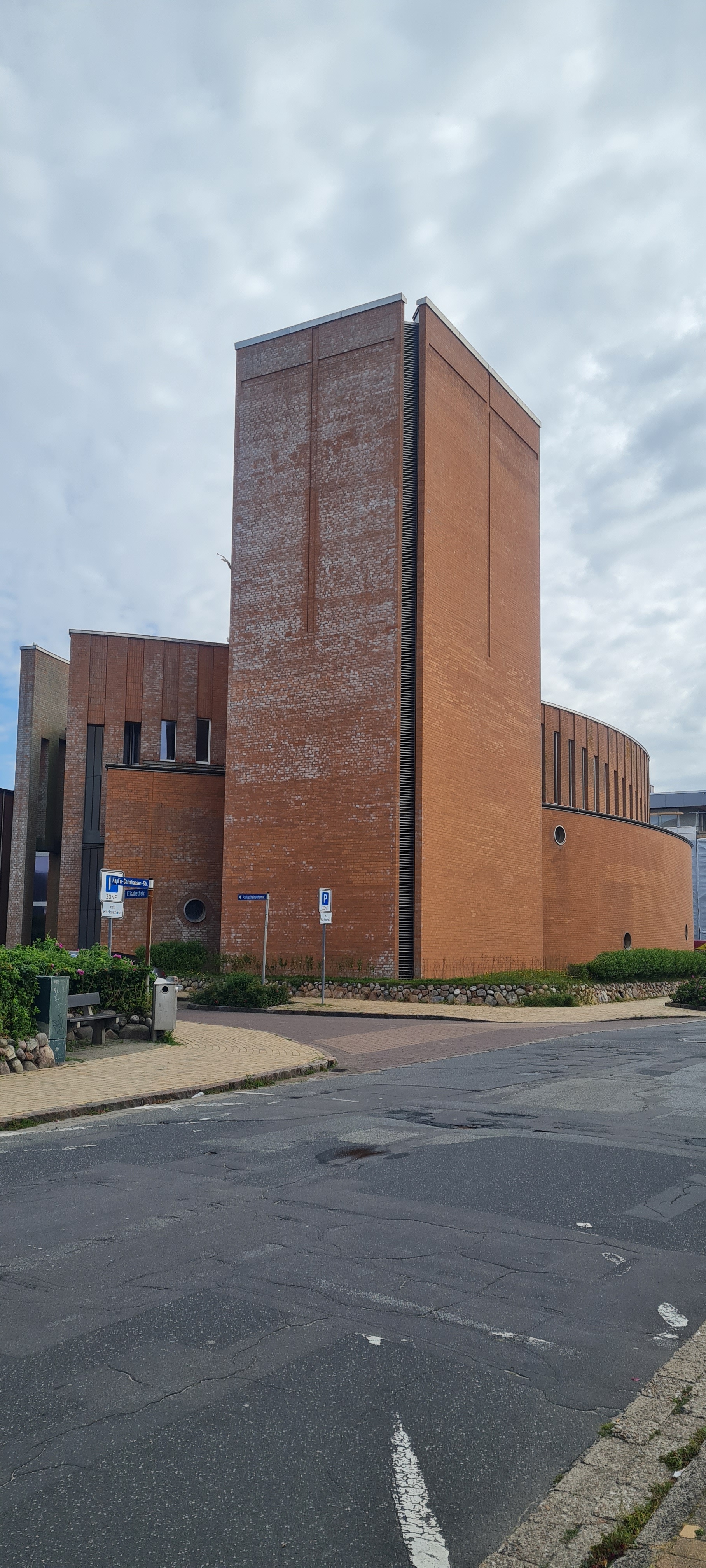 Es ist die Außenansicht der in roten Ziegelsteinen erbauten modern gestalteten Kirche mit eckigem Turm zu sehen