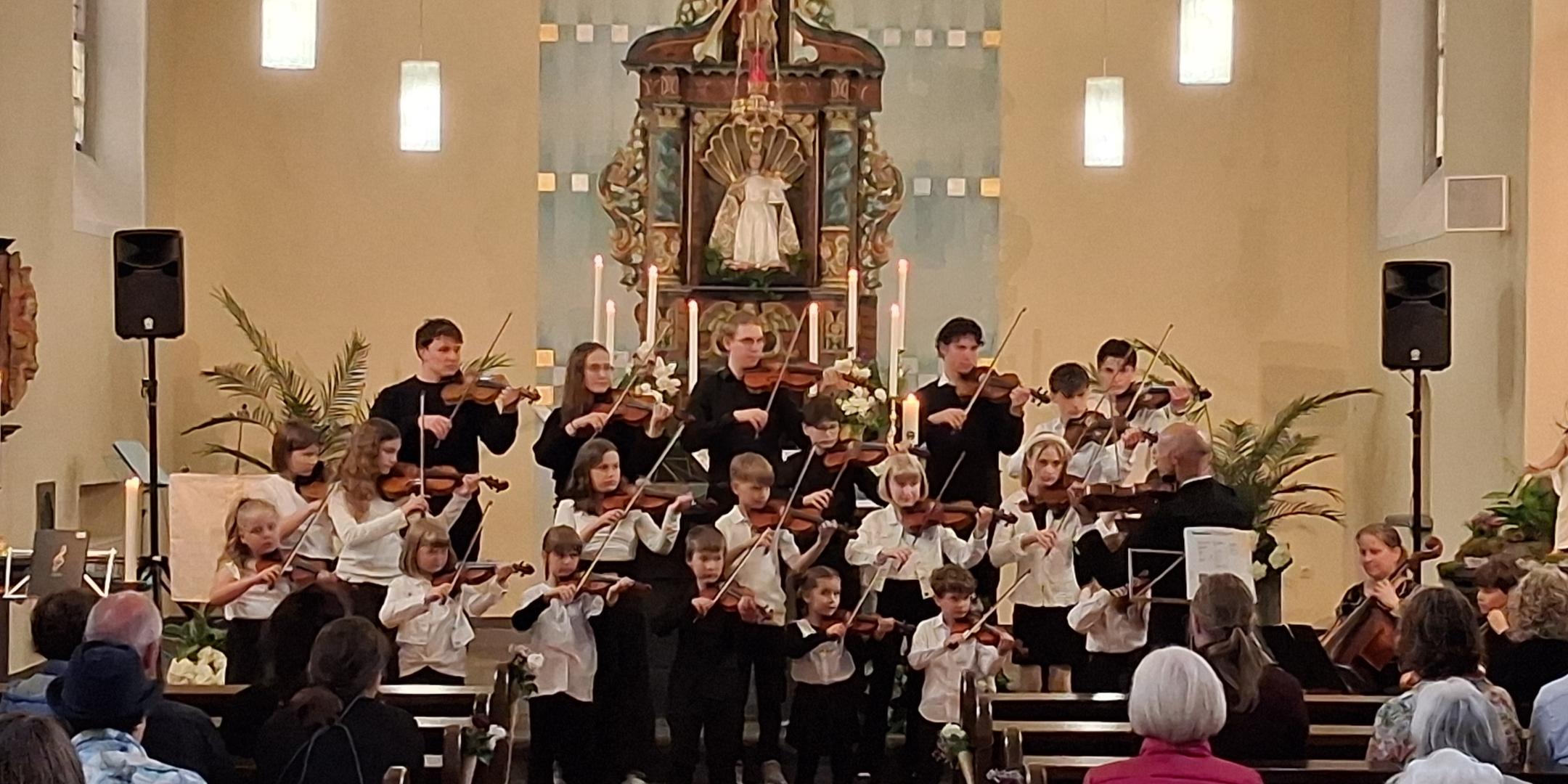 Man sieht den Altarraum mit Hochaltar der Kirche in Kalt, in dem ein Orchester aus Streichern, Kinder und Erwachsene stehen