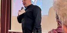 Man sieht einen Mann mit Mikrofon in der Hand, bei einem Vortrag, vor einer Leinwand stehen