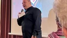 Man sieht einen Mann mit Mikrofon in der Hand, bei einem Vortrag, vor einer Leinwand stehen
