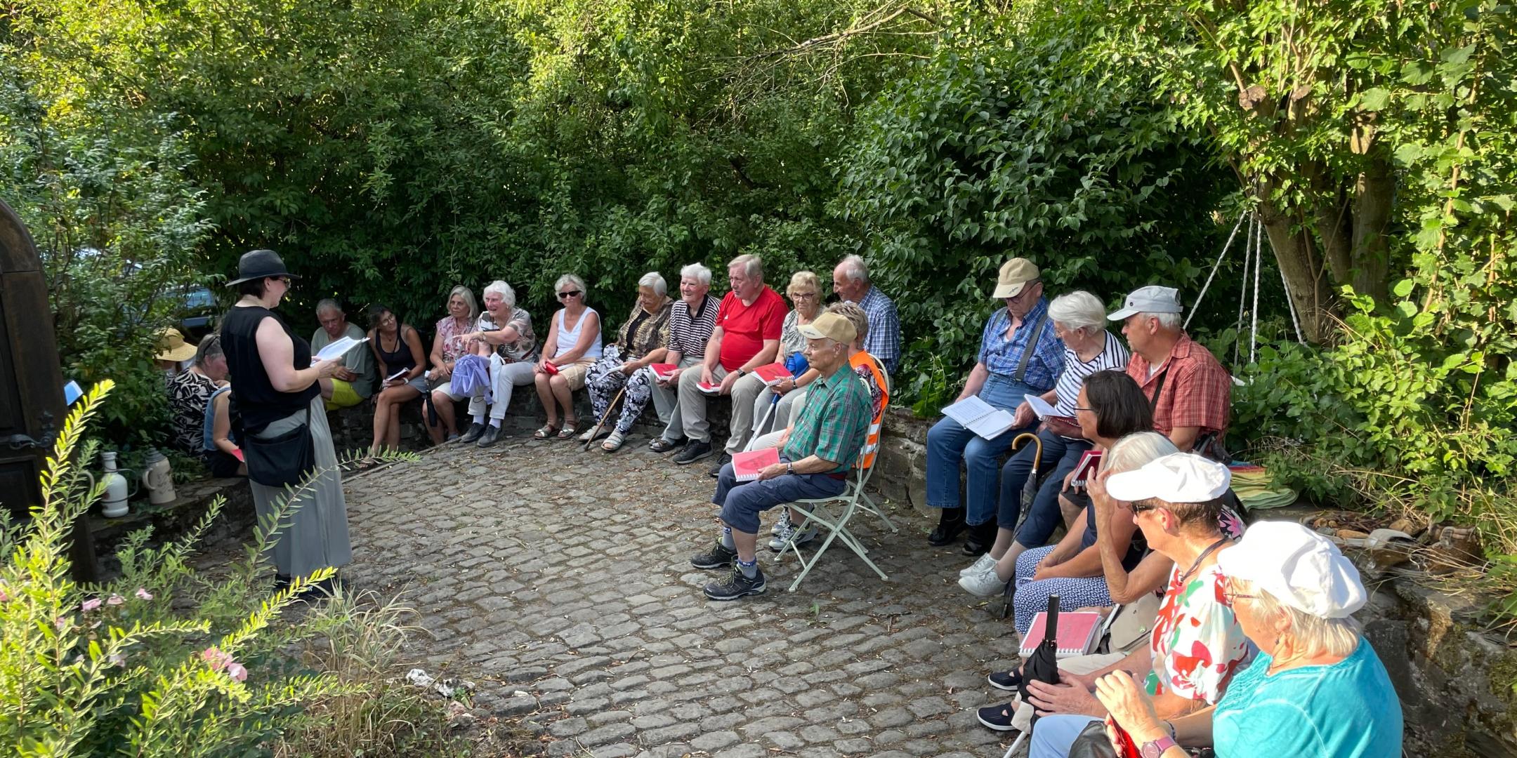 Man sieht Menschen auf Stühlen, die auf einer gepflasterten Fläche stehen, umringt vom grünen Sträuchern