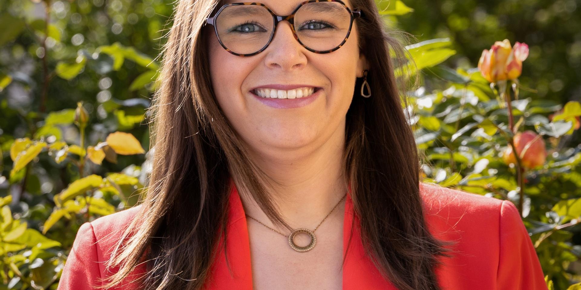 Man sieht eine lachende, junge Frau mit rotem Jackett über einer schwarzen Bluse vor einem frühlingshaften GArtenhintergrund