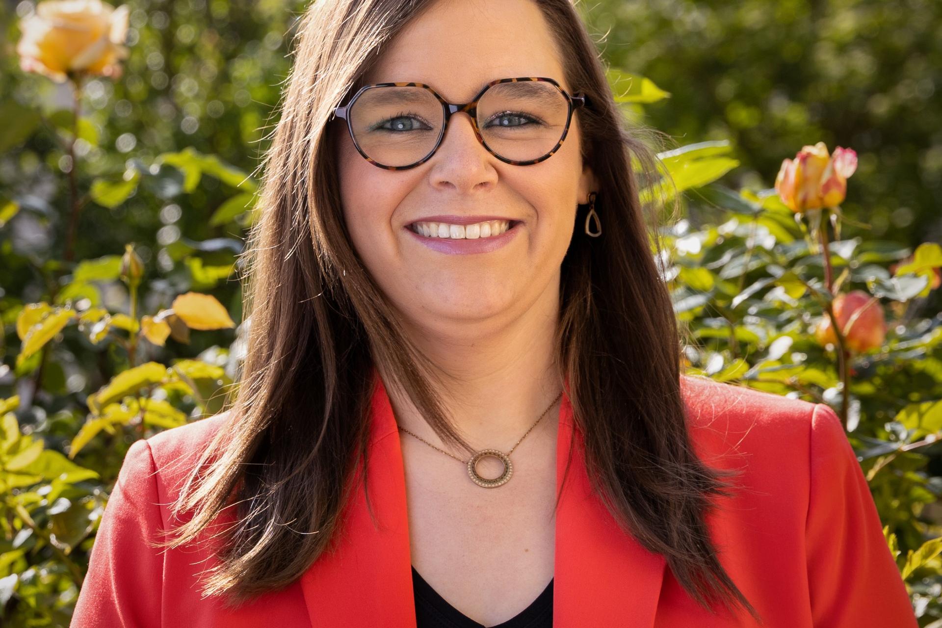 Man sieht eine lachende, junge Frau mit rotem Jackett über einer schwarzen Bluse vor einem frühlingshaften GArtenhintergrund