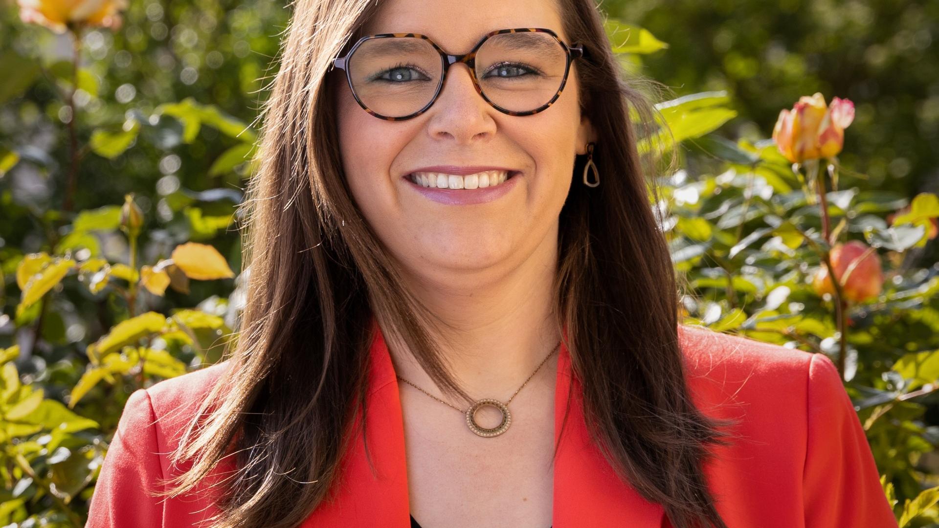 Man sieht eine lachende, junge Frau mit rotem Jackett über einer schwarzen Bluse vor einem frühlingshaften GArtenhintergrund