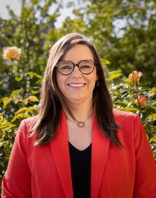 Man sieht eine lachende, junge Frau mit rotem Jackett über einer schwarzen Bluse vor einem frühlingshaften GArtenhintergrund