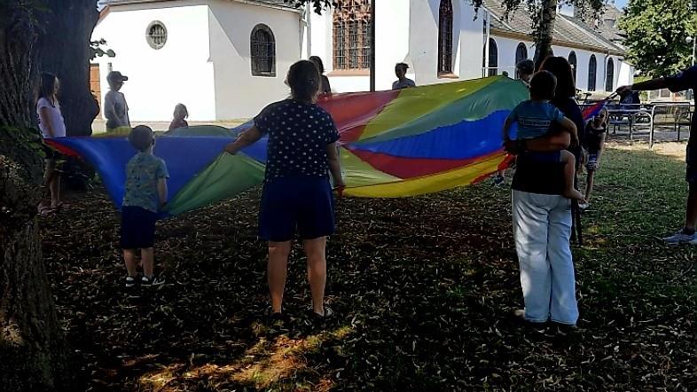 Es ist zu sehen wie vor der Mertlocher Kirche Kinder mit einem bunten Schwungtuch stehen