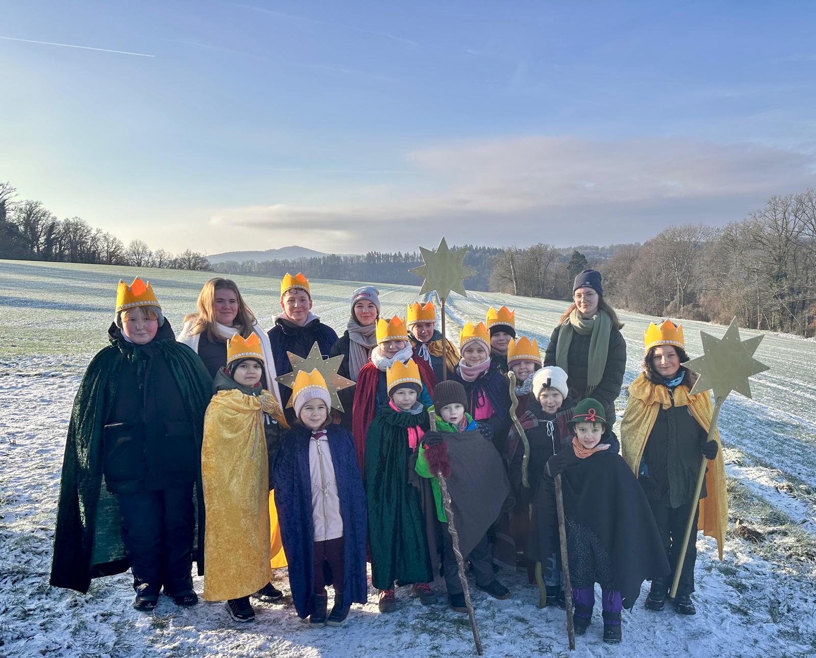 Es ist eine Gruppe Kinder zu sehen, die als Könige und Sternträger verkleidet auf einer schneebedeckten Wiese bei blauem Himmel fotografiert wurden
