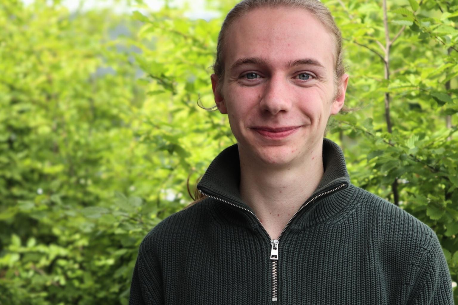 Man sieht einen gut gelaunten jungen Mann vor grünen Bäumen stehen