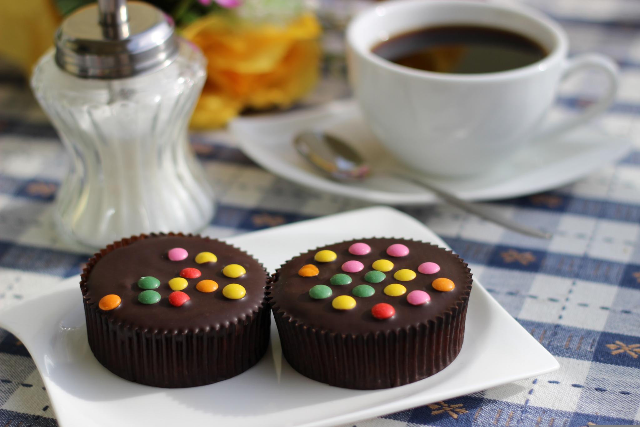Man sieht ein weißes Kaffeegedeck mit zwei von bunten Schokolinsen bedeckten Schokoladenörtchen