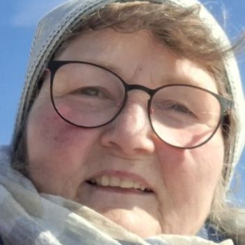 Man sieht das Proträt einer Frau mit helleren haaren, eine Mütze und Brille tragend