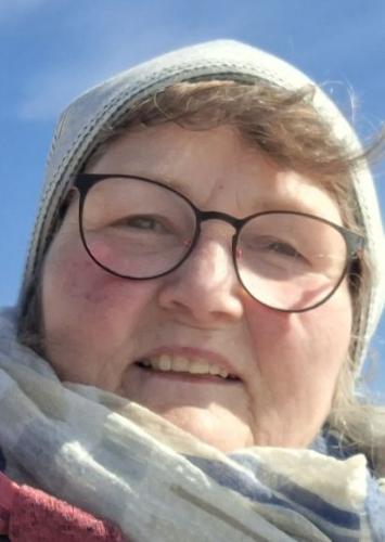 Man sieht das Proträt einer Frau mit helleren haaren, eine Mütze und Brille tragend